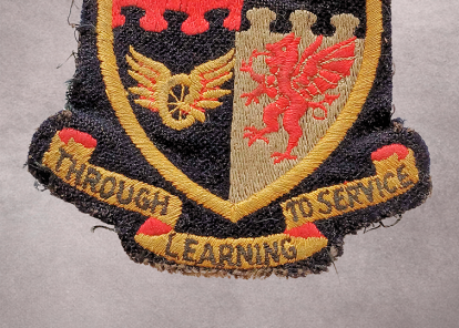 A worn, embroidered patch featuring a shield divided into four sections, each with distinct symbols, including an acorn and a lion. Below the shield, a banner reads 