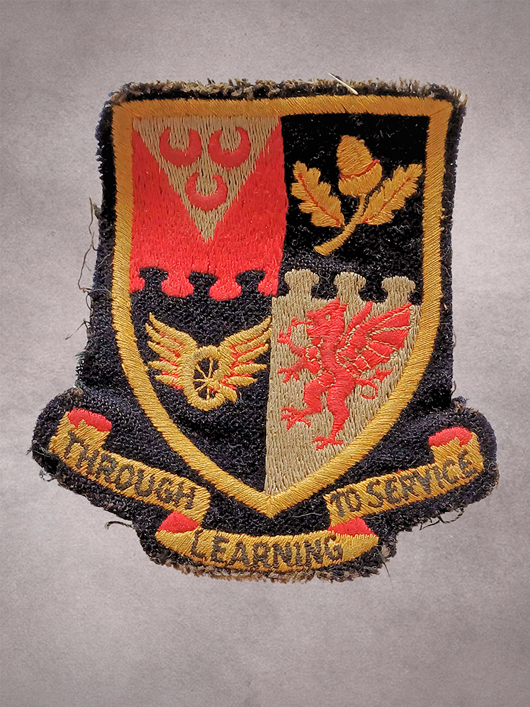 A worn, embroidered patch featuring a shield divided into four sections, each with distinct symbols, including an acorn and a lion. Below the shield, a banner reads 