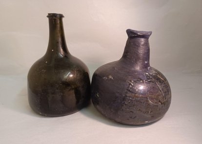 The image shows two ancient-looking glass bottles or jugs. One is a dark green bottle with an elongated neck and a round body. The other is a bulbous, dark gray ceramic jug with a short neck and rounded shape. The bottles appear to be antique or vintage in style, with a worn, weathered appearance.