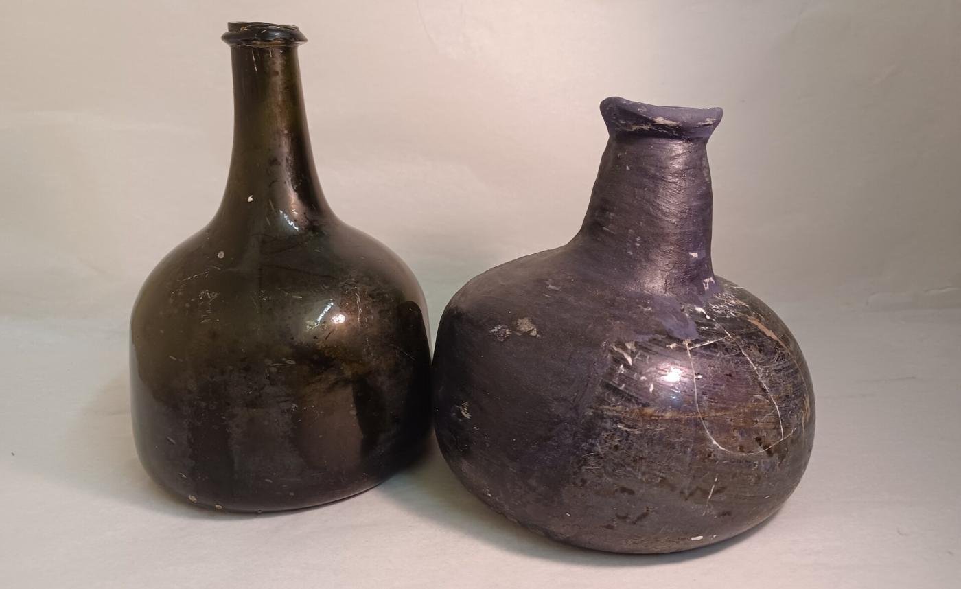 The image shows two ancient-looking glass bottles or jugs. One is a dark green bottle with an elongated neck and a round body. The other is a bulbous, dark gray ceramic jug with a short neck and rounded shape. The bottles appear to be antique or vintage in style, with a worn, weathered appearance.